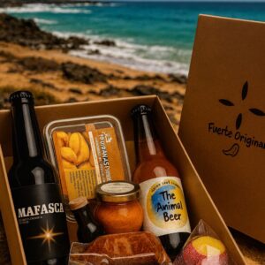 Caja de productos locales de Fuerteventura con cervezas artesanales, mojo rojo, puipanas, queso y dulces, fotografiada en la costa rocosa con el mar turquesa de fondo.