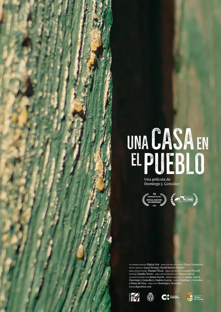 Cartel de la película “Una casa en el pueblo” sobre textura de madera verde, con el título en blanco y laureles de selección de festival.